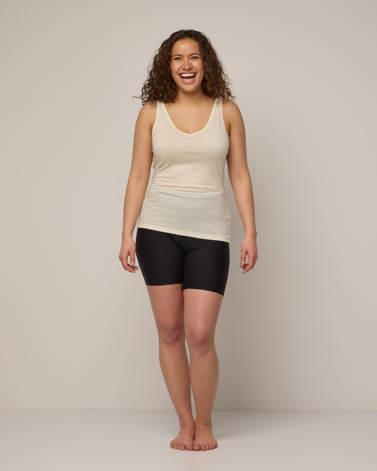 Model wearing Essential Layering Tank in Natural White, an off-white cream color. Essential Layering Tank is a merino wool sleeveless, reversible, slim fit top available in sizes XS-3X.