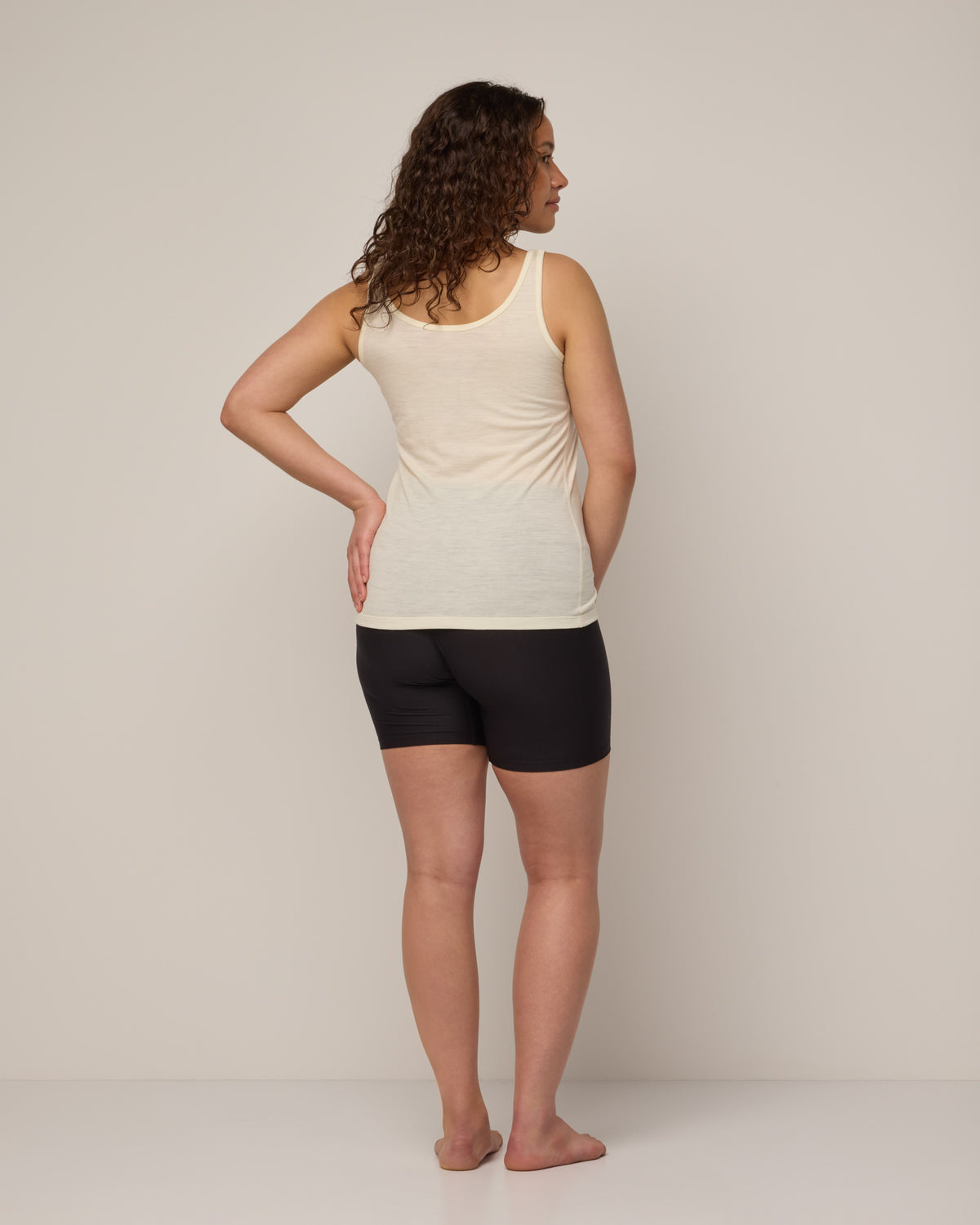 Model wearing Essential Layering Tank in Natural White, an off-white cream color. Essential Layering Tank is a merino wool sleeveless, reversible, slim fit top available in sizes XS-3X.