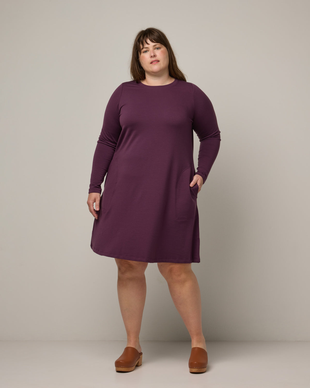 Model wearing Eliza Swing Dress in Marionberry, a dark berry purple color. Eliza Swing Dress is a merino wool long sleeve, crew neck, knee length swing dress with pockets available in sizes XS-3X Regular, XS-3X Long.