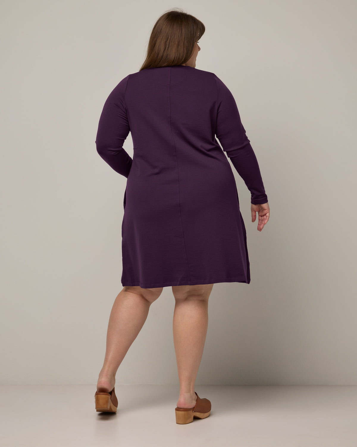 Model wearing Eliza Swing Dress in Cassis, a dark purple color. Eliza Swing Dress is a merino wool long sleeve, crew neck, knee length swing dress with pockets available in sizes XS-3X Regular, XS-3X Long.