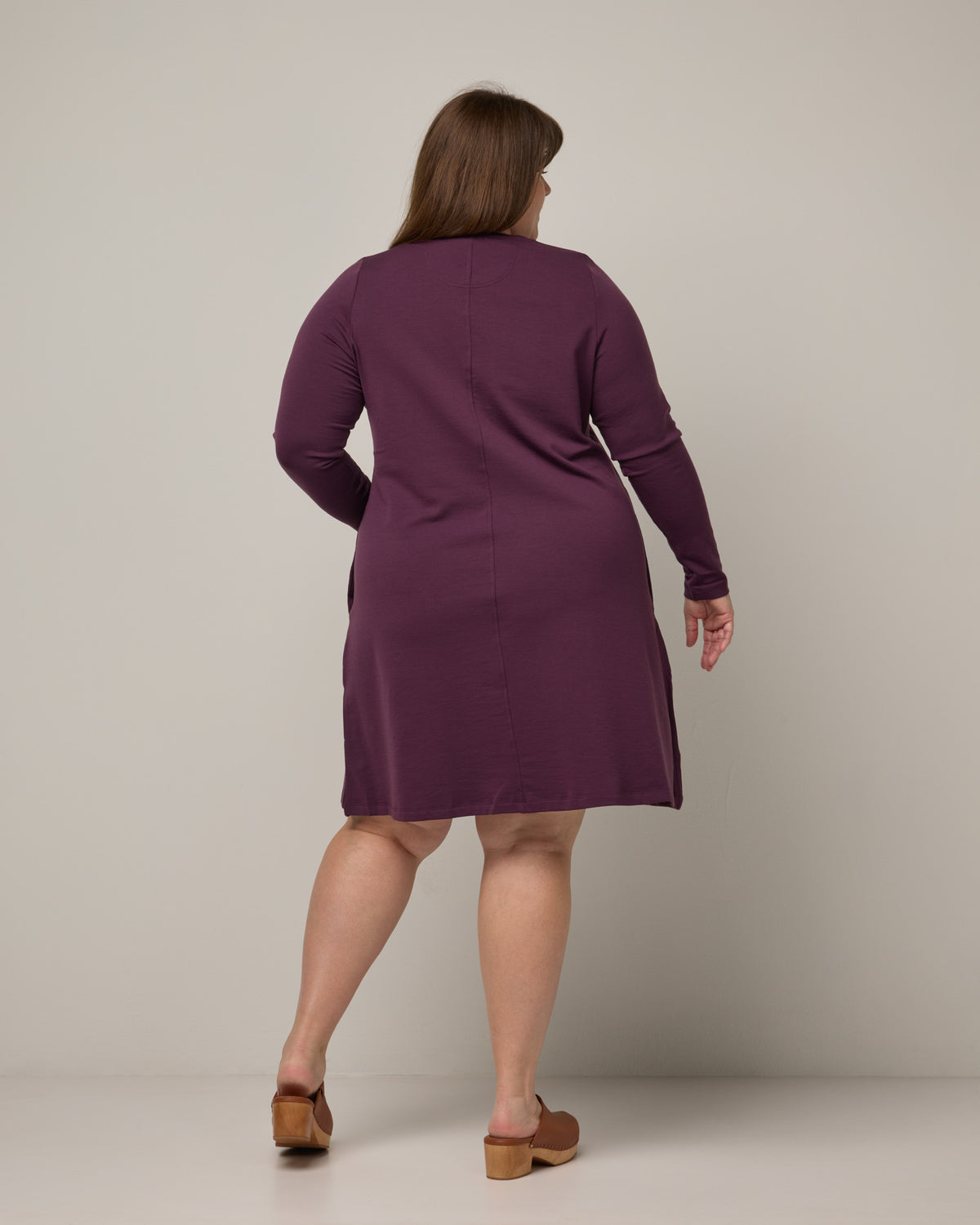 Model wearing Eliza Swing Dress in Marionberry, a dark berry purple color. Eliza Swing Dress is a merino wool long sleeve, crew neck, knee length swing dress with pockets available in sizes XS-3X Regular, XS-3X Long.