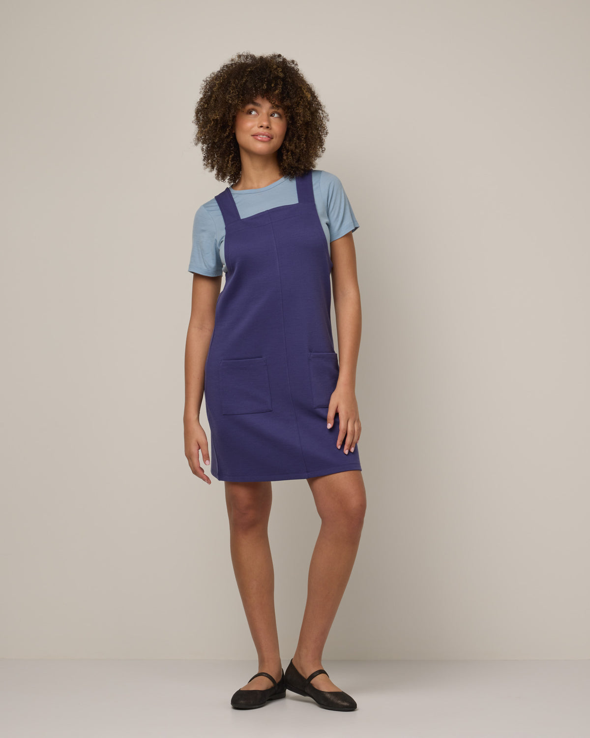 Model wearing Goldie Pinafore Dress in Iris Blue, a vibrant purple-blue color. Goldie Pinafore Dress is a merino wool sleeveless, square neck, knee length shift dress with pockets available in sizes XS-3X.
