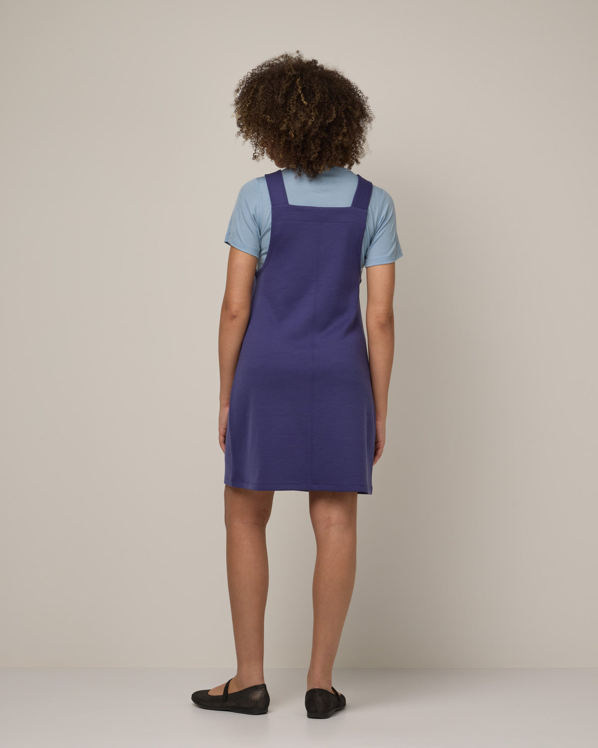 Model wearing Goldie Pinafore Dress in Iris Blue, a vibrant purple-blue color. Goldie Pinafore Dress is a merino wool sleeveless, square neck, knee length shift dress with pockets available in sizes XS-3X.