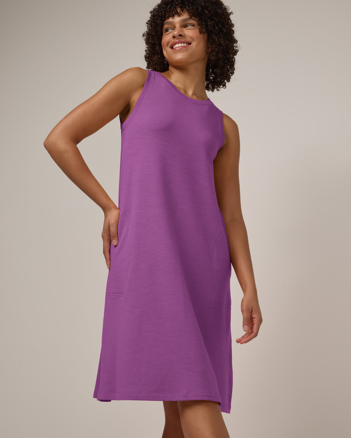 Model wearing Sierra Tank Dress in Purple Pansy, a bright, saturated purple color. Sierra Tank Dress is a merino wool sleeveless, crew neck, knee length swing dress with pockets available in sizes XS-3X Regular, XS-3X Long, XS-XL Petite.
