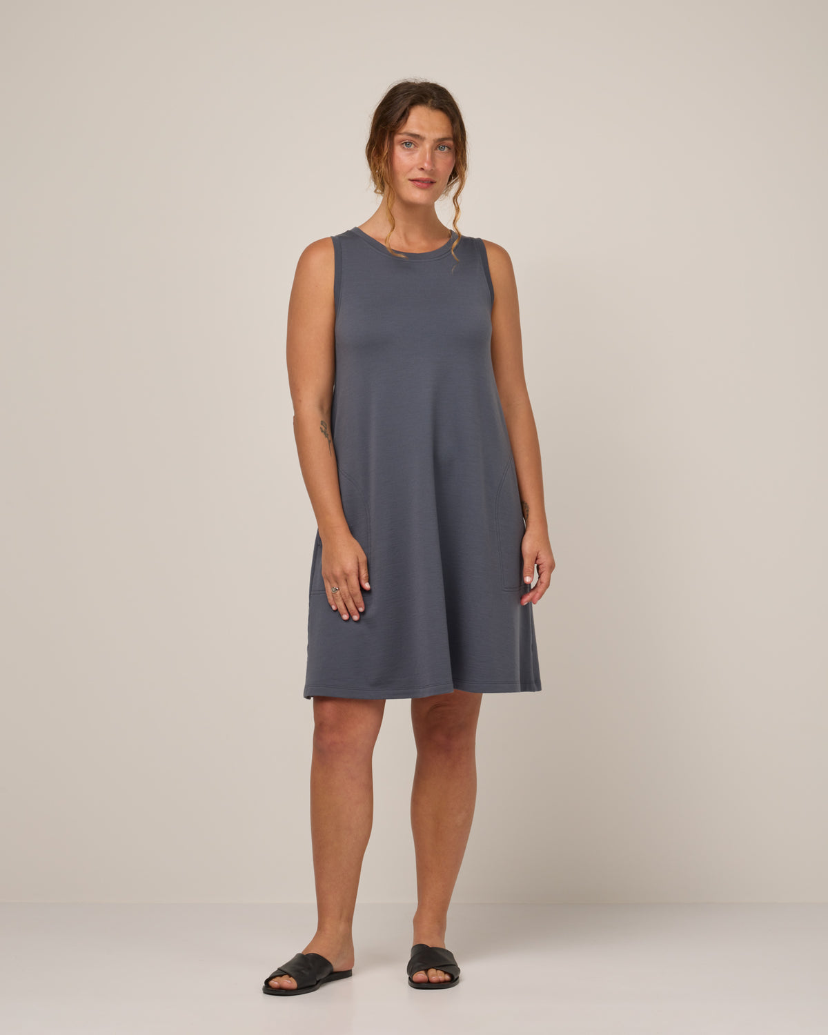 Model wearing Sierra Tank Dress in Odyssey Gray, a neutral purplish-gray color. Sierra Tank Dress is a merino wool sleeveless, crew neck, knee length swing dress with pockets available in sizes XS-3X Regular, XS-3X Long.