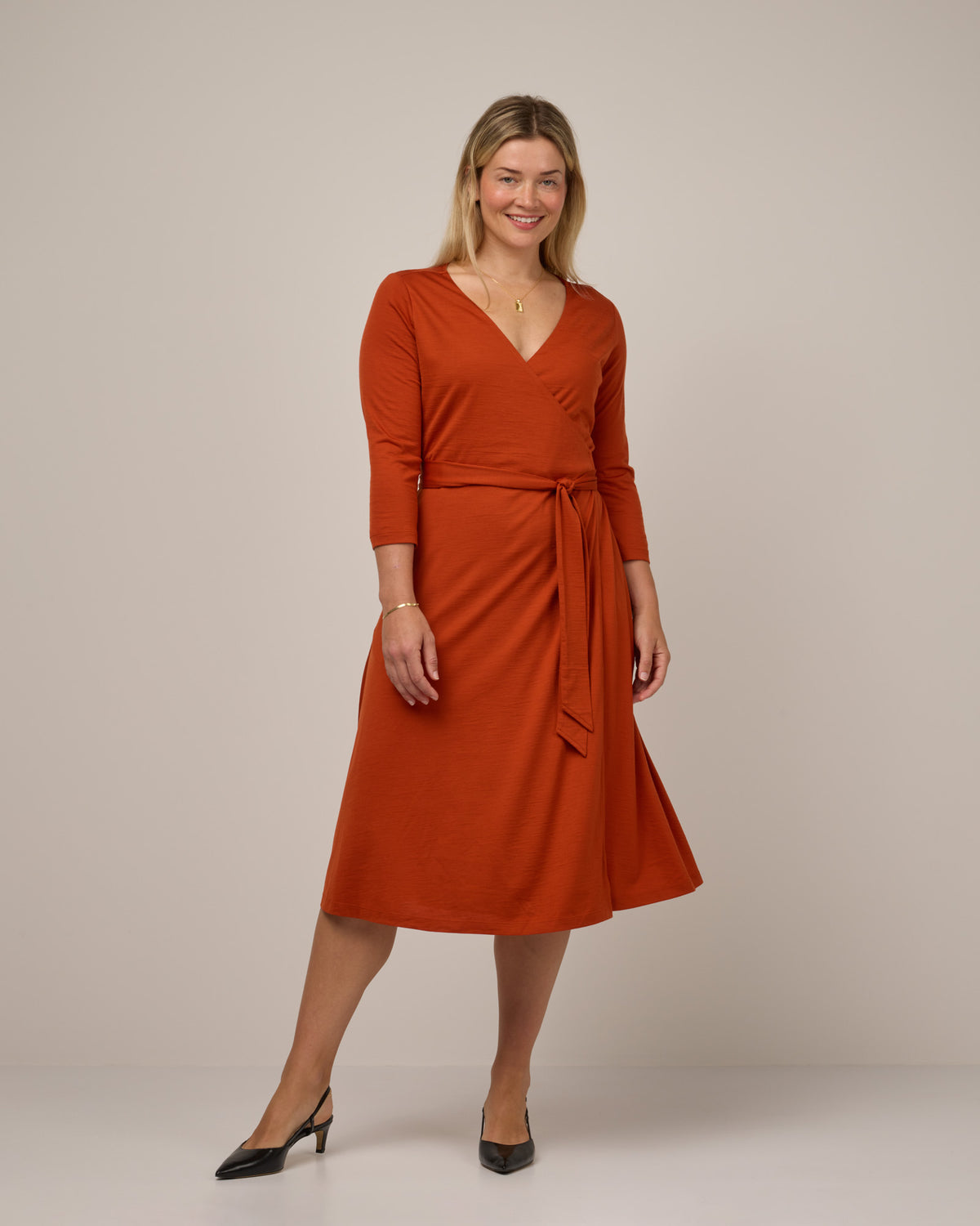Model wearing Alana Wrap Dress in Terracotta, a saturated red-orange color. Alana Wrap Dress is a merino wool long sleeve, v-neck, midi fit & flare dress with pockets available in sizes XS-3X.