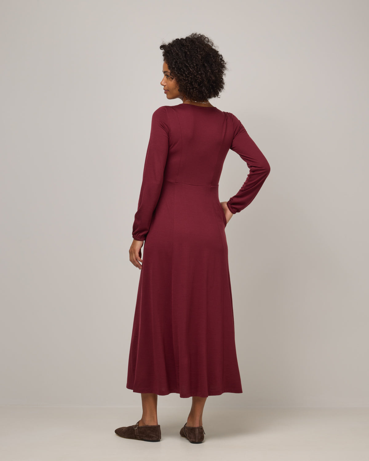 Model wearing Ahna Empire Waist Dress in Burgundy. Ahna Empire Waist Dress is a merino wool long sleeve, square neck, maxi fit & flare dress with pockets available in sizes XS-3X.