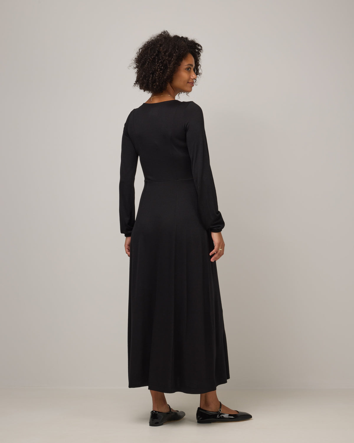 Model wearing Ahna Empire Waist Dress in Black. Ahna Empire Waist Dress is a merino wool long sleeve, square neck, maxi fit & flare dress with pockets available in sizes XS-3X.