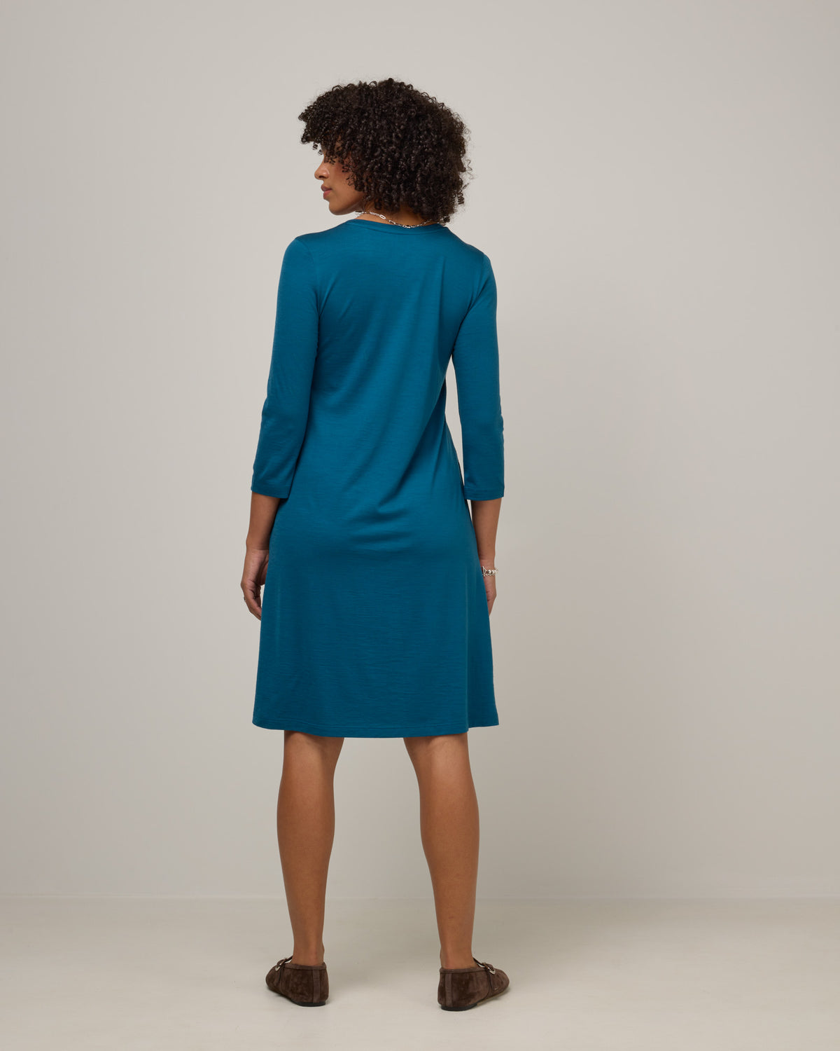 Model wearing Willow Swing Dress in Ocean Teal, a medium blue-green color. Willow Swing Dress is a merino wool long sleeve, v-neck, knee length swing dress with pockets available in sizes XS-3X Regular, XS-3X Long.