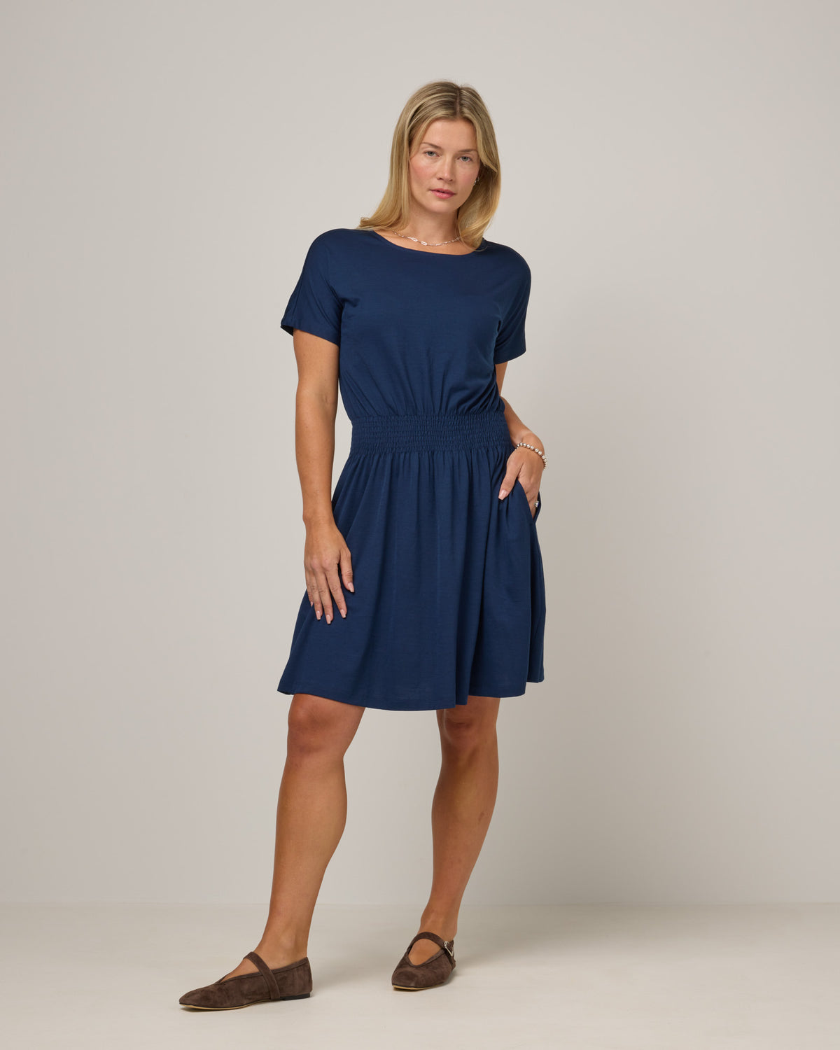 Model wearing Norma Smocked Waist Dress in Marine Blue, a deep royal blue color. Norma Smocked Waist Dress is a merino wool short sleeve, crew neck, knee length fit & flare dress with pockets available in sizes XS-3X.