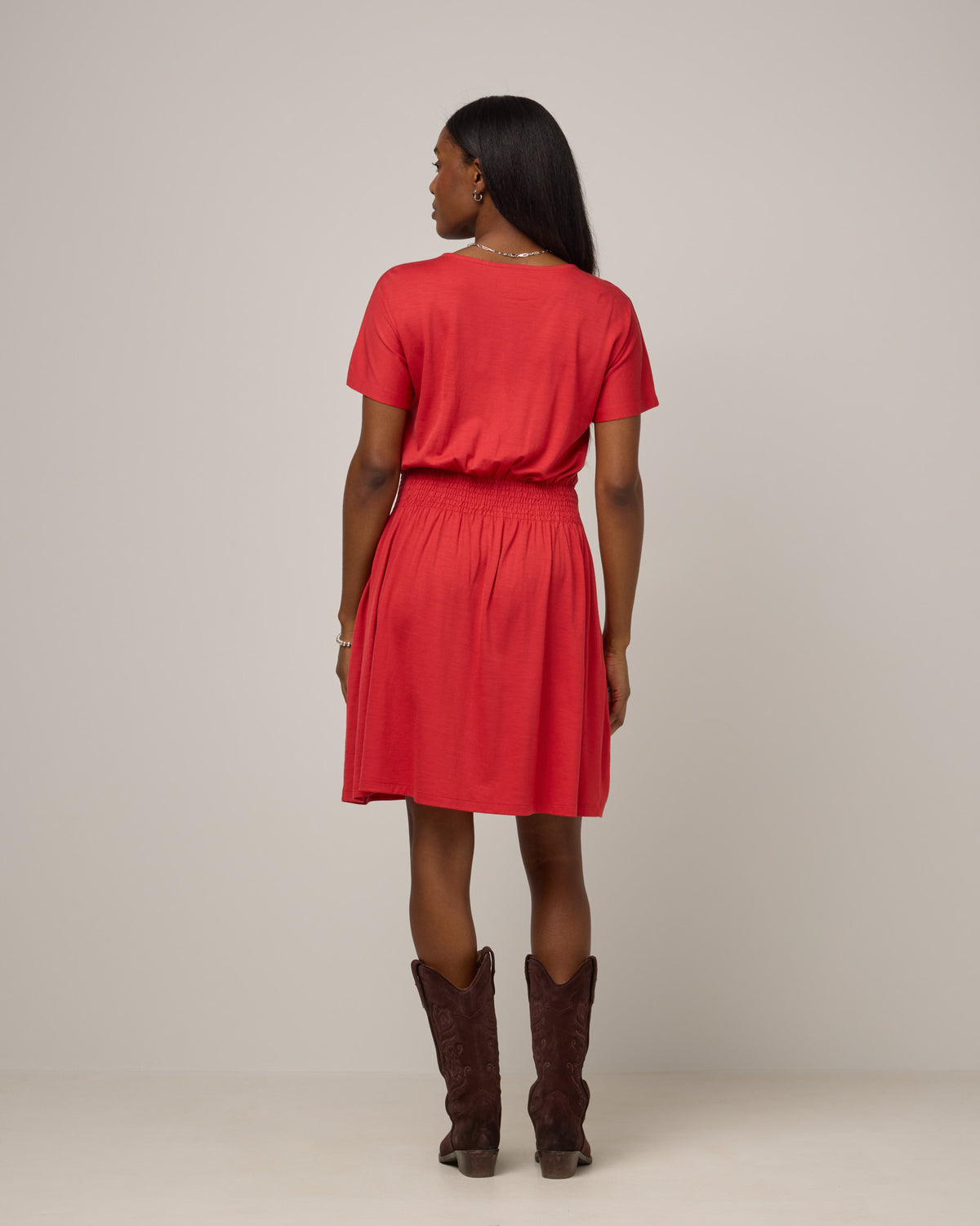 Model wearing Norma Smocked Waist Dress in Fruit Punch, a vivid red-orange color. Norma Smocked Waist Dress is a merino wool short sleeve, crew neck, knee length fit & flare dress with pockets available in sizes XS-3X.