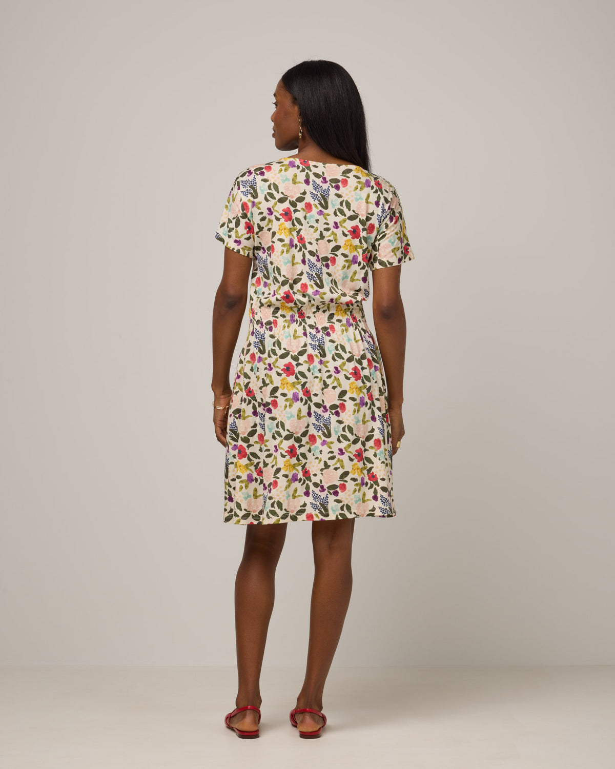 Model wearing Norma Smocked Waist Dress in Watercolor Floral Print, a multi-colored floral print on a cream background. Norma Smocked Waist Dress is a merino wool short sleeve, crew neck, knee length fit & flare dress with pockets available in sizes XS-3X.