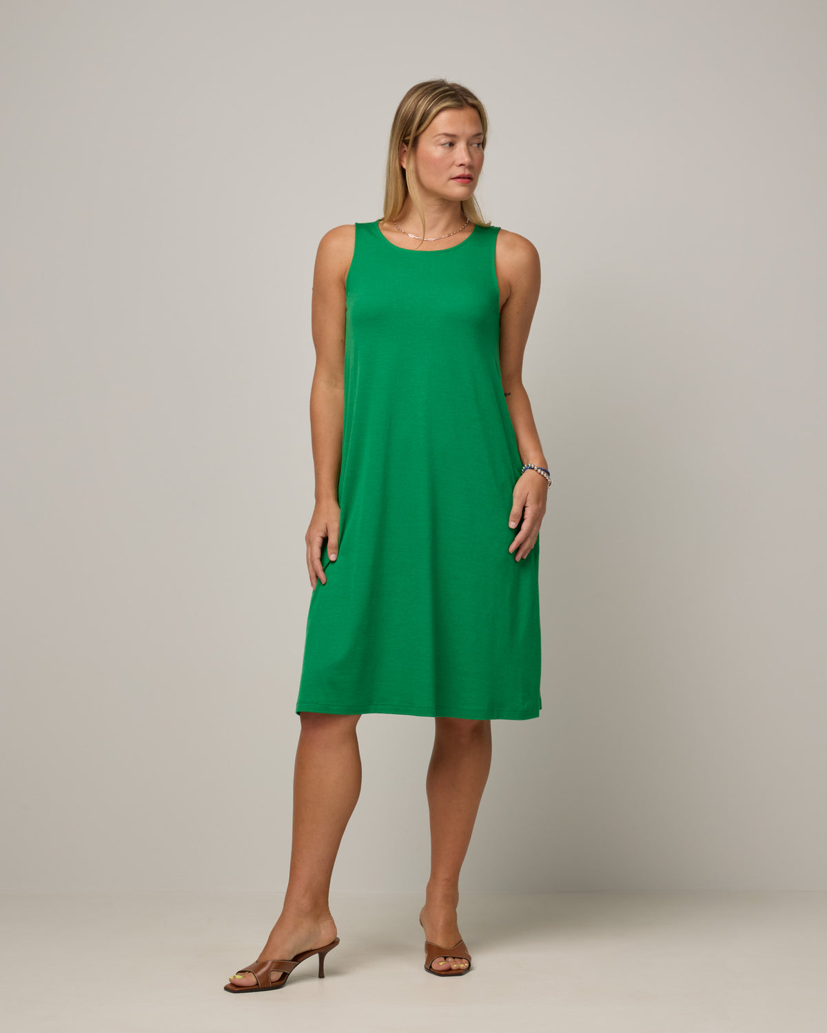 Model wearing Camellia Tank Dress in Verde, a bright Kelly green color. Camellia Tank Dress is a merino wool sleeveless, crew neck, knee length swing dress with pockets available in sizes XS-3X Regular, XS-3X Long.