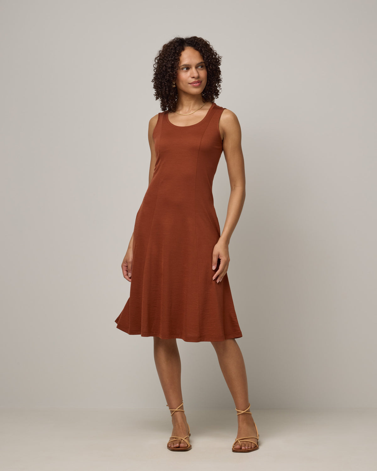 Model wearing Flora Swing Dress in Cinnamon, a reddish-brown rust color. Flora Swing Dress is a merino wool sleeveless, scoop neck, knee length swing dress with pockets available in sizes XS-3X.