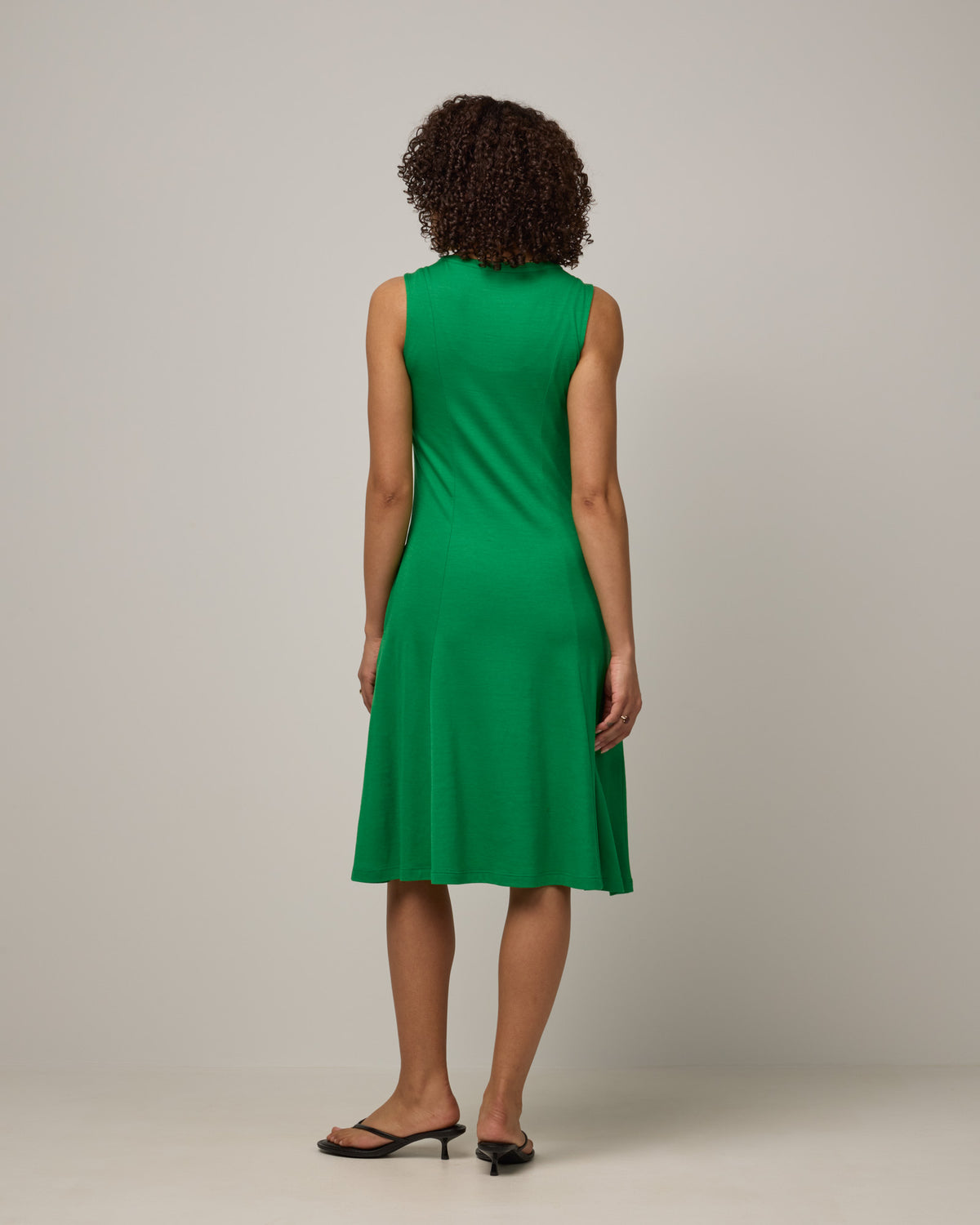 Model wearing Flora Swing Dress in Verde, a bright Kelly green color. Flora Swing Dress is a merino wool sleeveless, scoop neck, knee length swing dress with pockets available in sizes XS-3X.