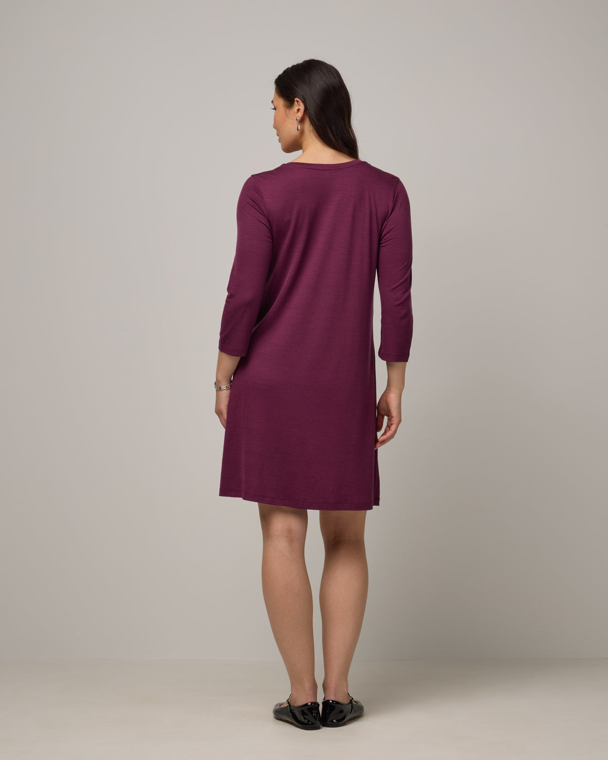 Model wearing Willow Swing Dress in Plum, a reddish-purple color. Willow Swing Dress is a merino wool long sleeve, v-neck, knee length swing dress with pockets available in sizes XS-3X Regular, XS-3X Long.