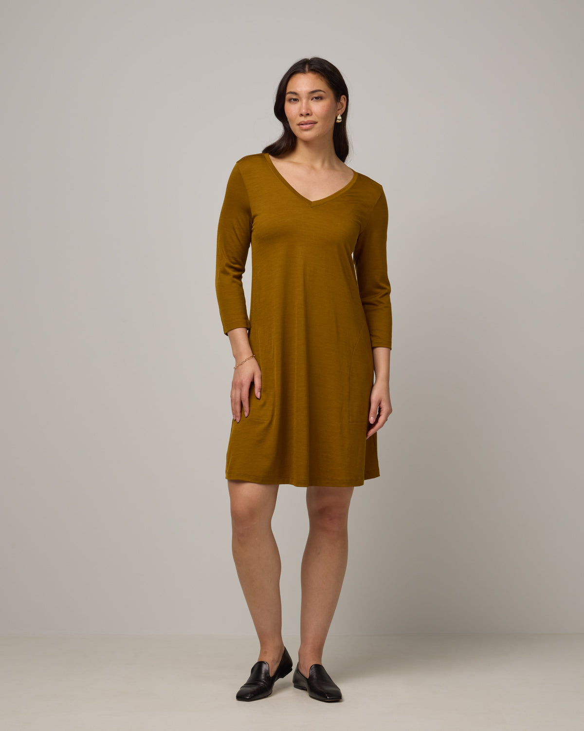 Model wearing Willow Swing Dress in Spiced Pear, a dark mustard color. Willow Swing Dress is a merino wool long sleeve, v-neck, knee length swing dress with pockets available in sizes XS-3X Regular, XS-3X Long.