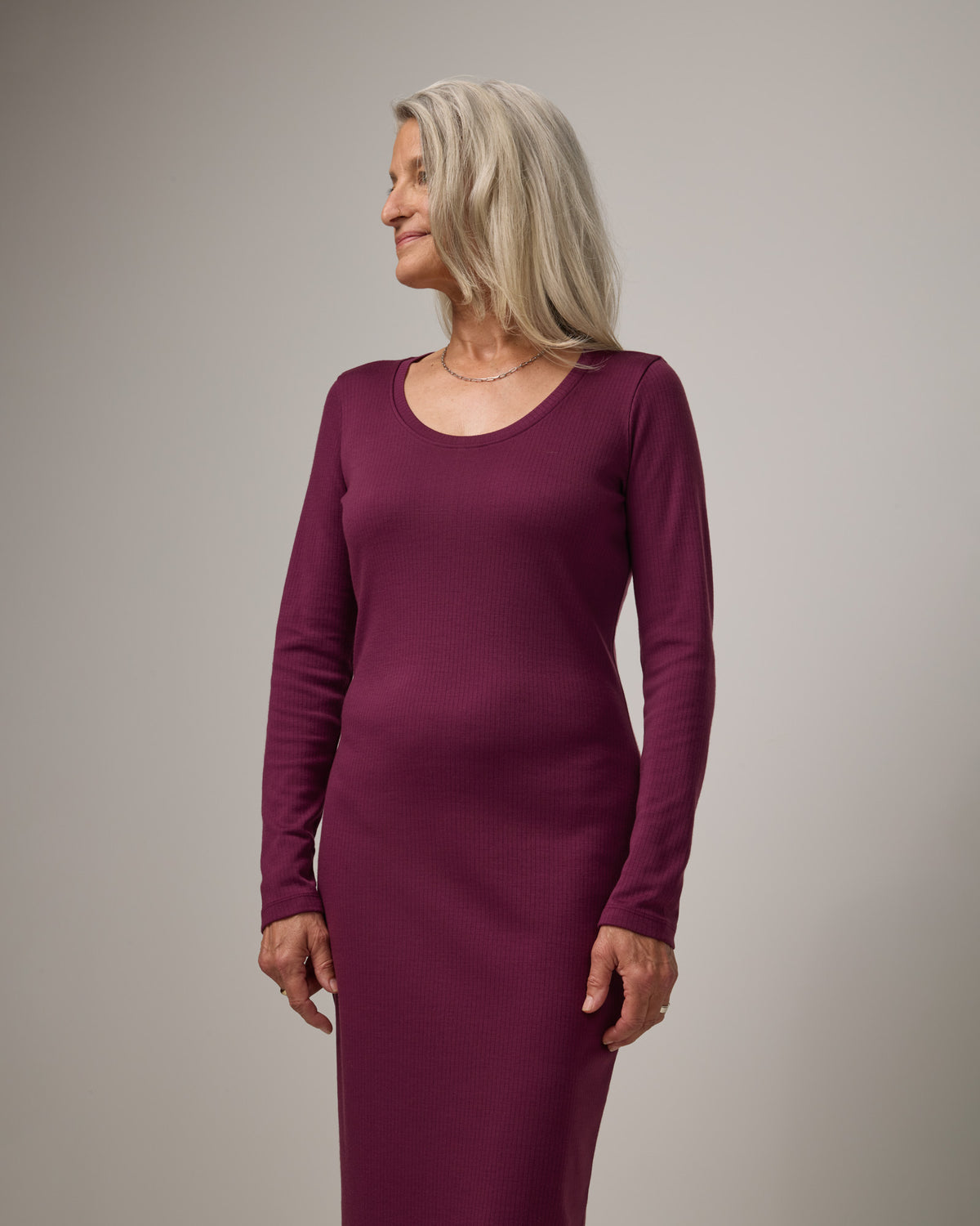 Model wearing Lex Ribbed Dress in Plum, a reddish-purple color. Lex Ribbed Dress is a merino wool long sleeve, scoop neck, fitted midi dress available in sizes XS-XXL Regular & XS-XL Petite.