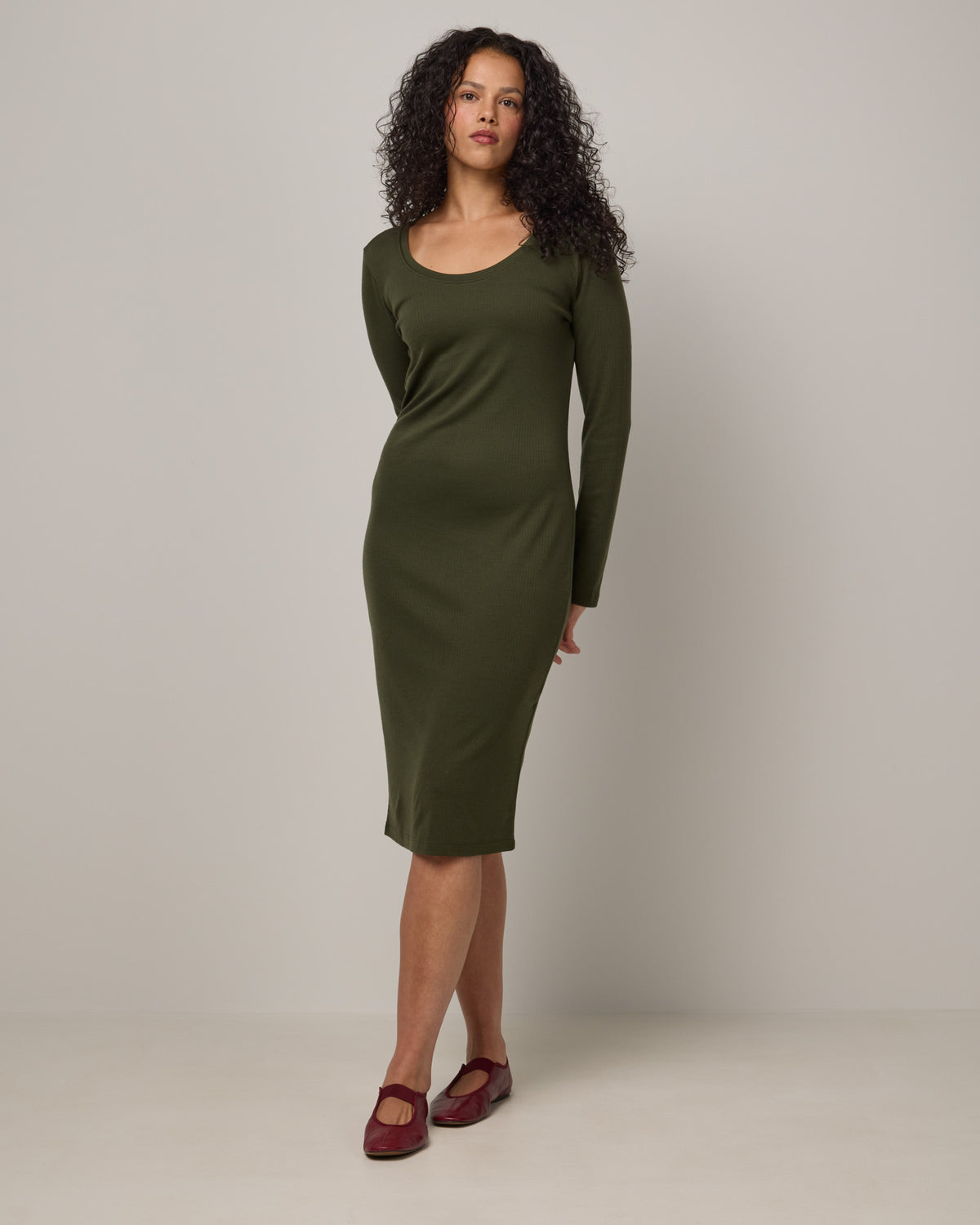 Model wearing Lex Ribbed Dress in Forest Night, a dark forest green color. Lex Ribbed Dress is a merino wool long sleeve, scoop neck, fitted midi dress available in sizes XS-XXL Regular & XS-XL Petite.