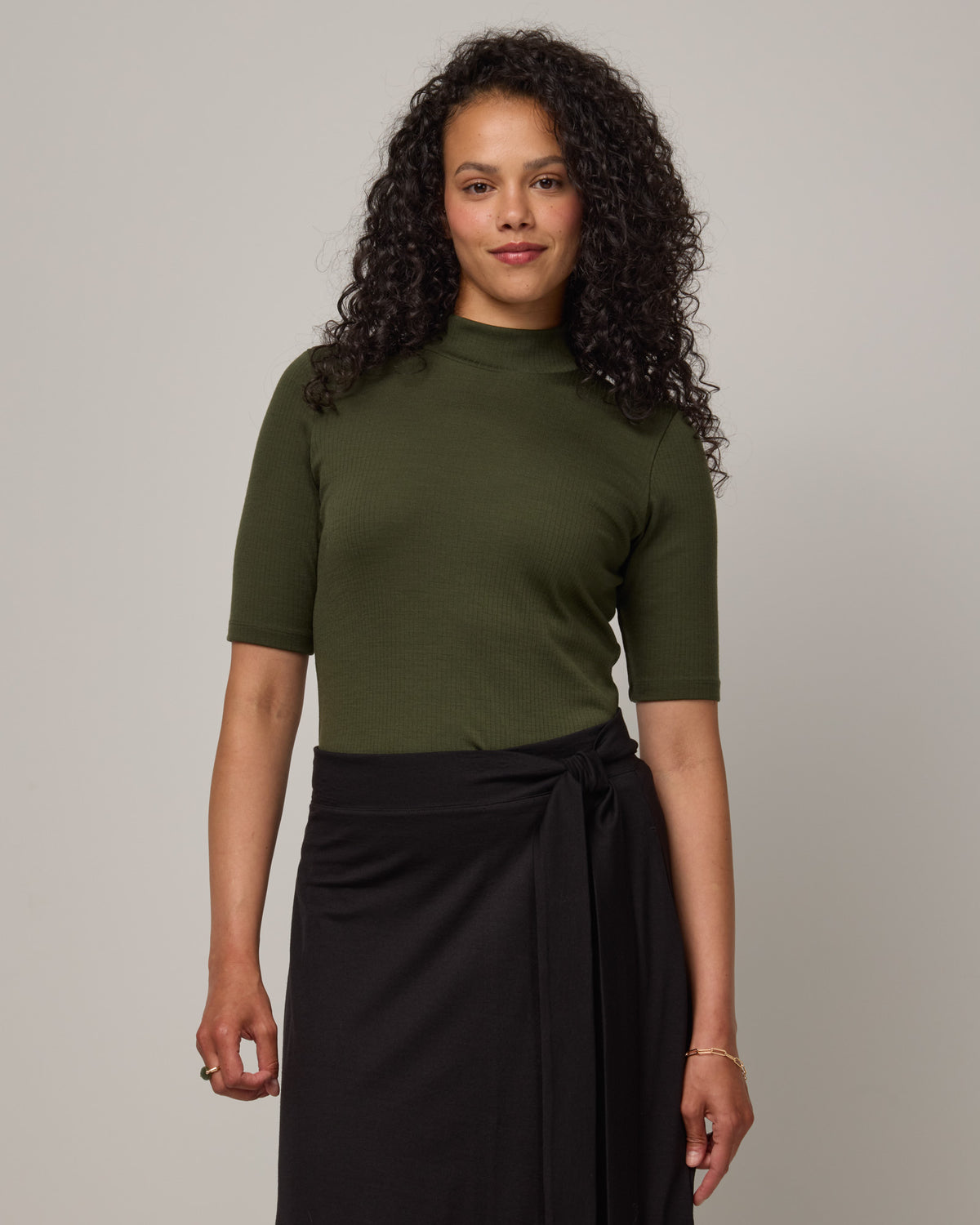 Model wearing Krystal Mock Neck Top in Forest Night, a dark forest green color. Krystal Mock Neck Top is a merino wool half-sleeve, mock-neck ribbed tee available in sizes XS-XXL.