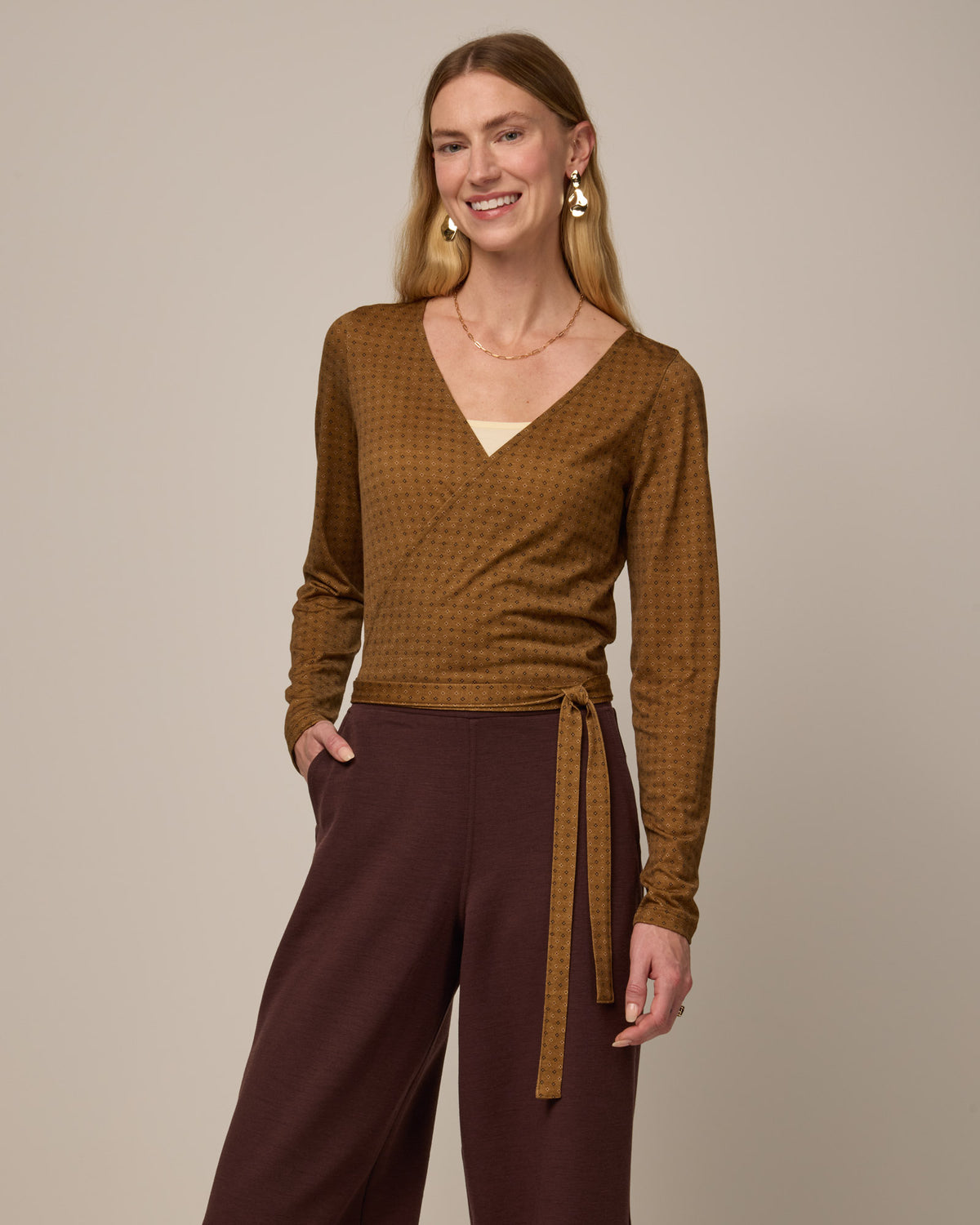 Model wearing Natalia Wrap Top in Geo Stamp Print, a dainty, neutral, geometric print on a dark ochre background. Natalia Wrap Top is a merino wool long sleeve, v-neck, slim fit top available in sizes XS-3X.