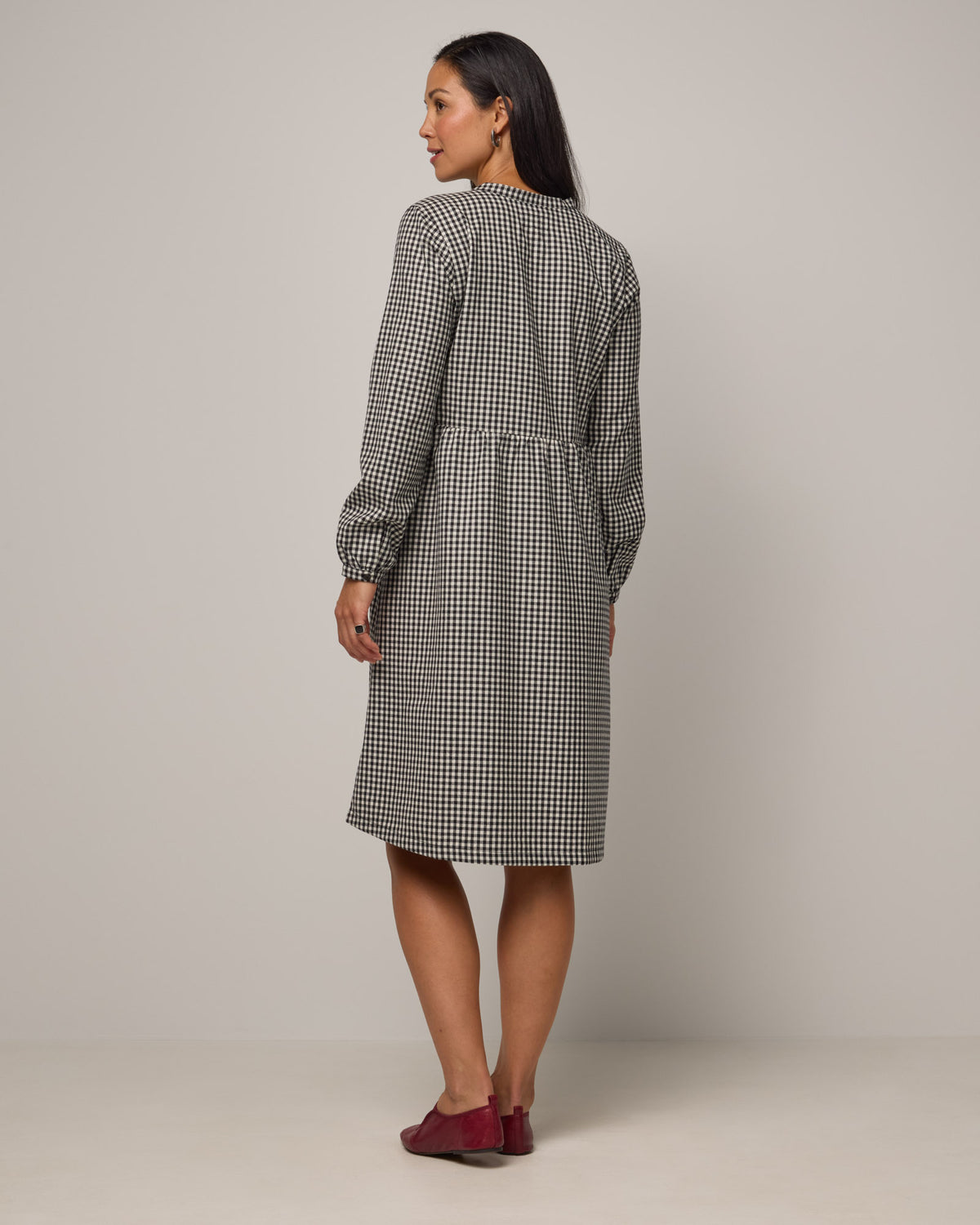 Model wearing Marika Brushed Twill Dress in Black & White Gingham. Marika Brushed Twill Dress is a merino wool long sleeve, gathered, midi swing dress with a button placket available in sizes XS-XXL.