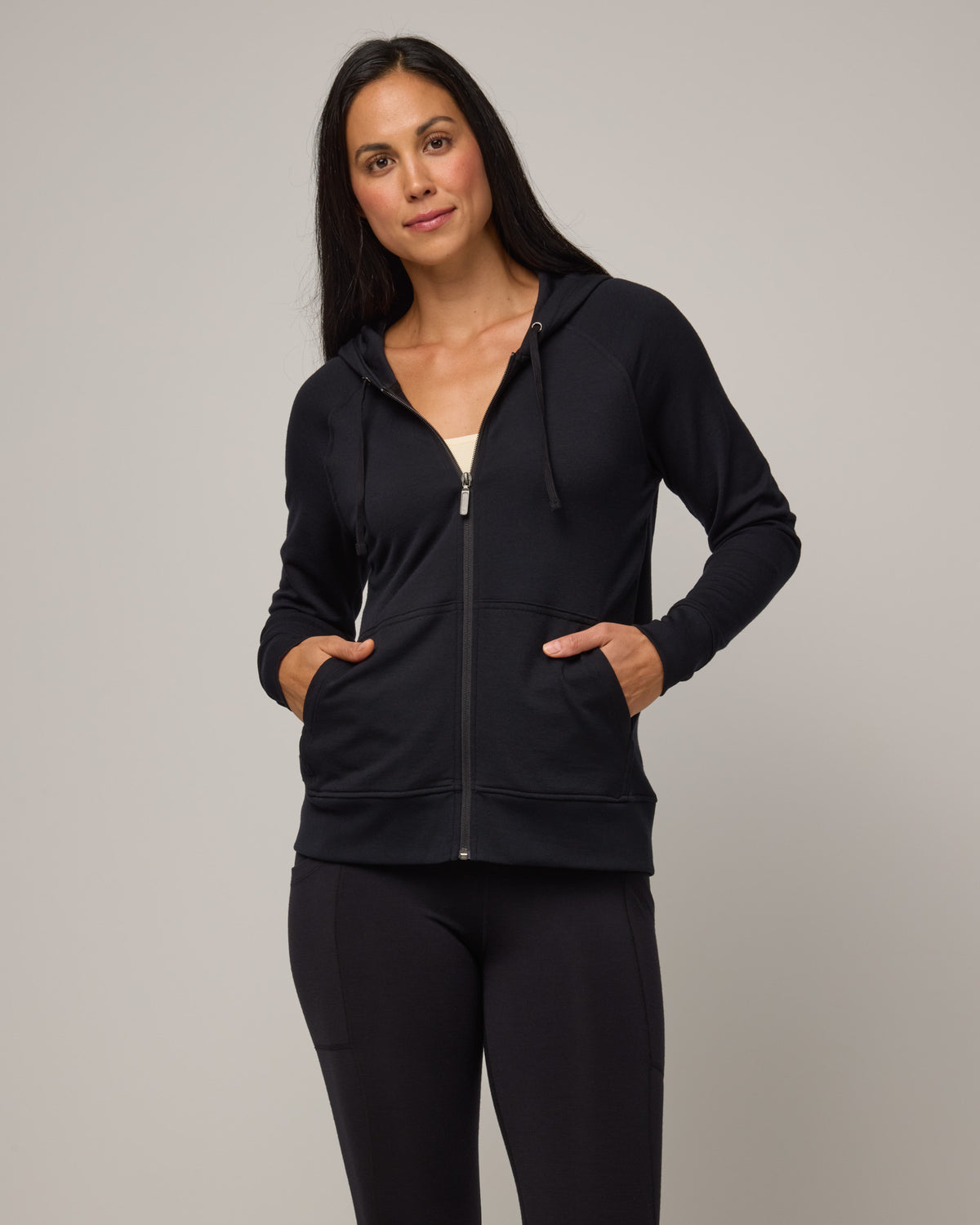 Model wearing Aspen Full-Zip Hoodie in Black. Aspen Full-Zip Hoodie is a merino wool long sleevezip hoodie with pockets available in sizes XS-3X.