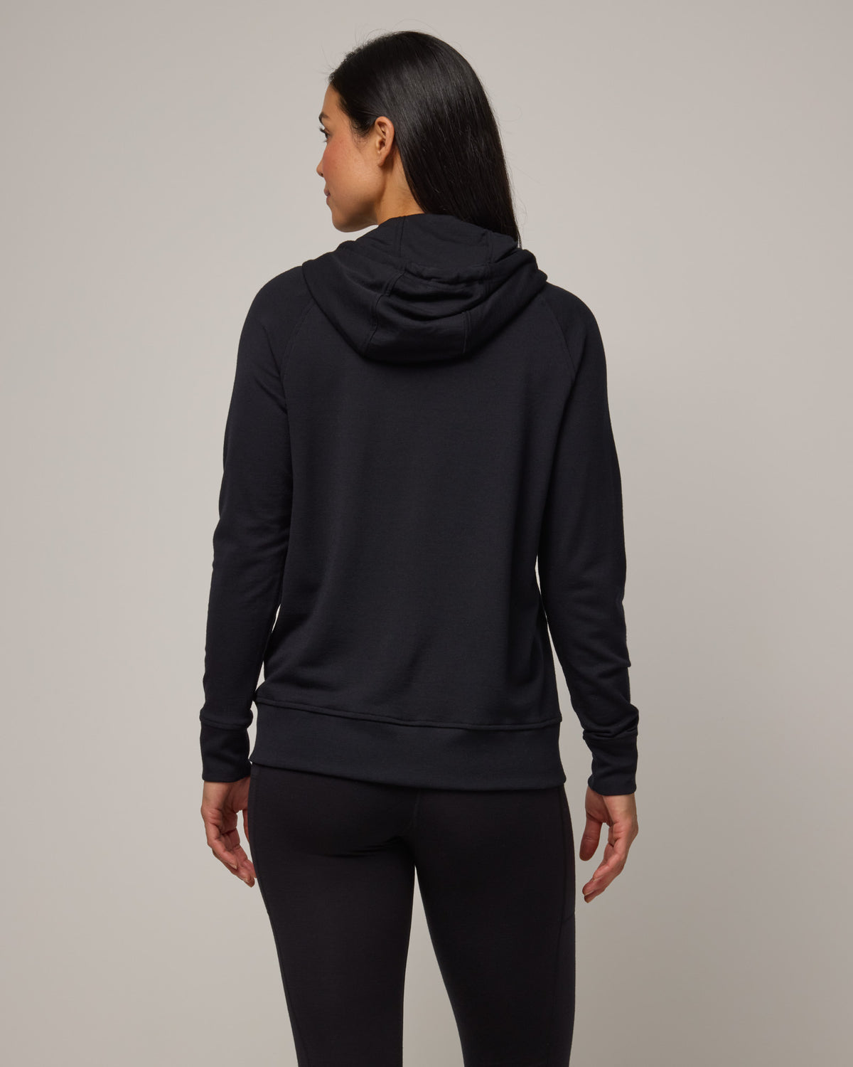 Model wearing Aspen Full-Zip Hoodie in Black. Aspen Full-Zip Hoodie is a merino wool long sleevezip hoodie with pockets available in sizes XS-3X.