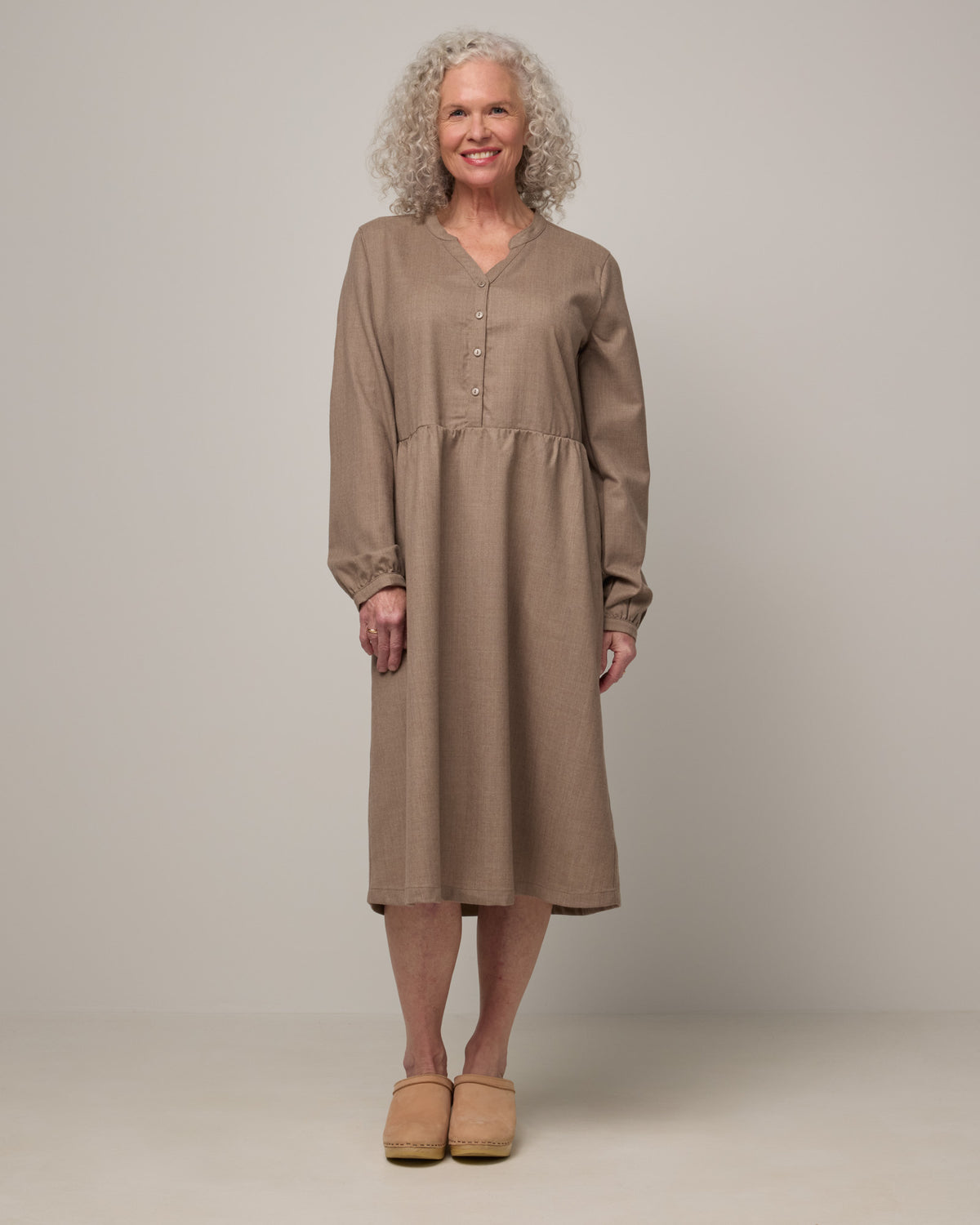 Model wearing Marika Brushed Twill Dress in Taupe heather, a heathered tan color. Marika Brushed Twill Dress is a merino wool long sleeve, gathered, midi swing dress with a button placket available in sizes XS-XXL.