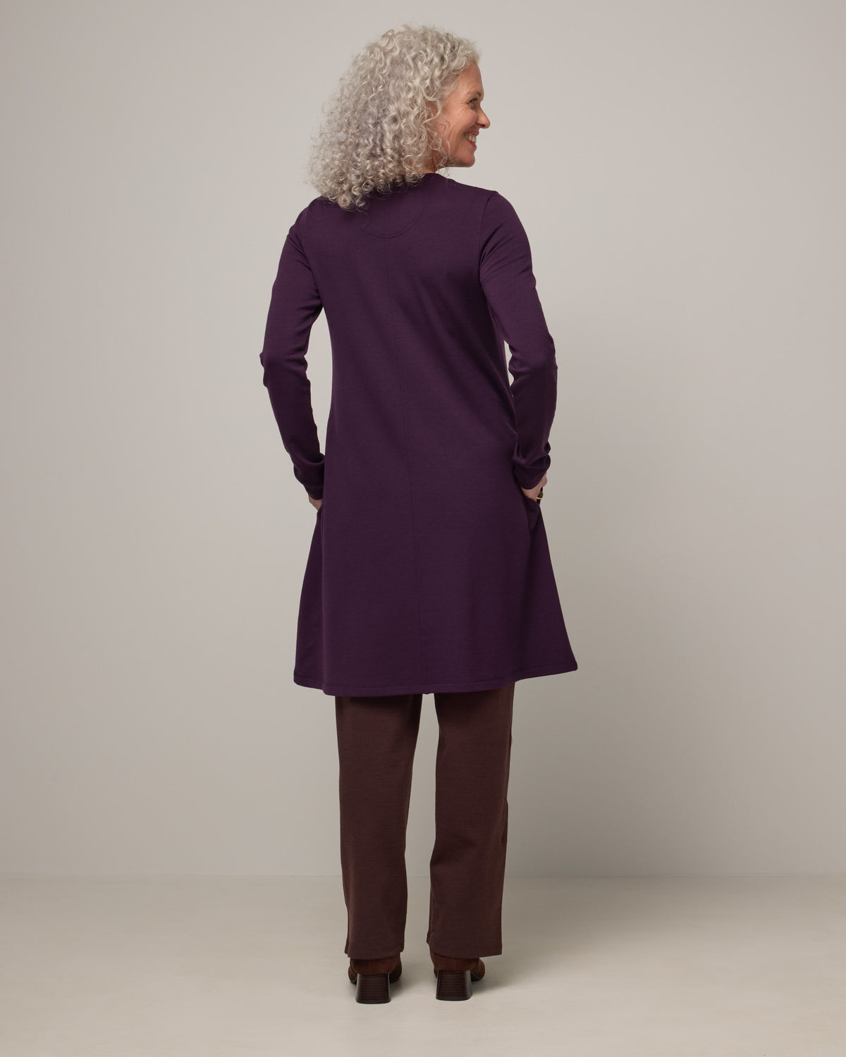 Model wearing Eliza Swing Dress in Cassis, a dark purple color. Eliza Swing Dress is a merino wool long sleeve, crew neck, knee length swing dress with pockets available in sizes XS-3X Regular, XS-3X Long.