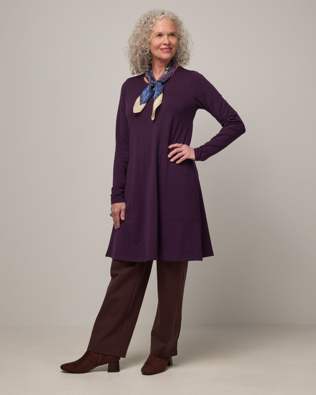 Model wearing Eliza Swing Dress in Cassis, a dark purple color. Eliza Swing Dress is a merino wool long sleeve, crew neck, knee length swing dress with pockets available in sizes XS-3X Regular, XS-3X Long.