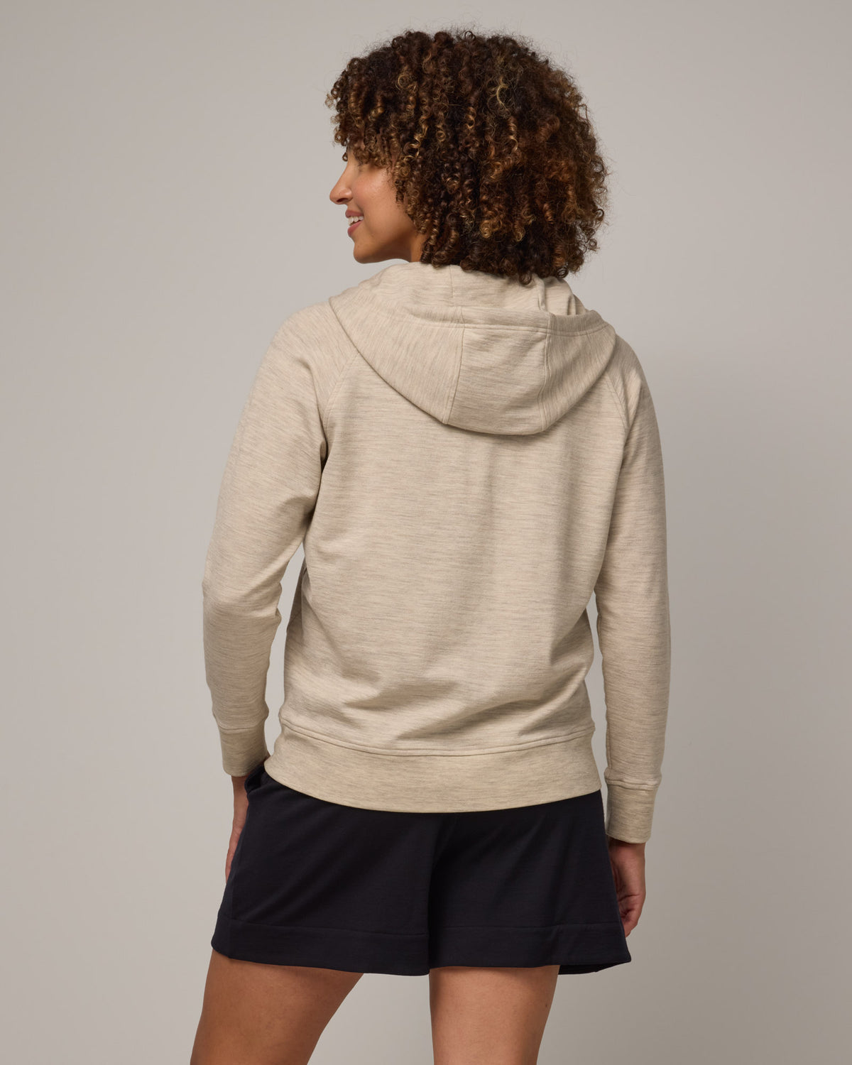 Model wearing Aspen Full-Zip Hoodie in Oatmeal Heather, a cream heather color. Aspen Full-Zip Hoodie is a merino wool long sleeve zip hoodie with pockets available in sizes XS-3X.